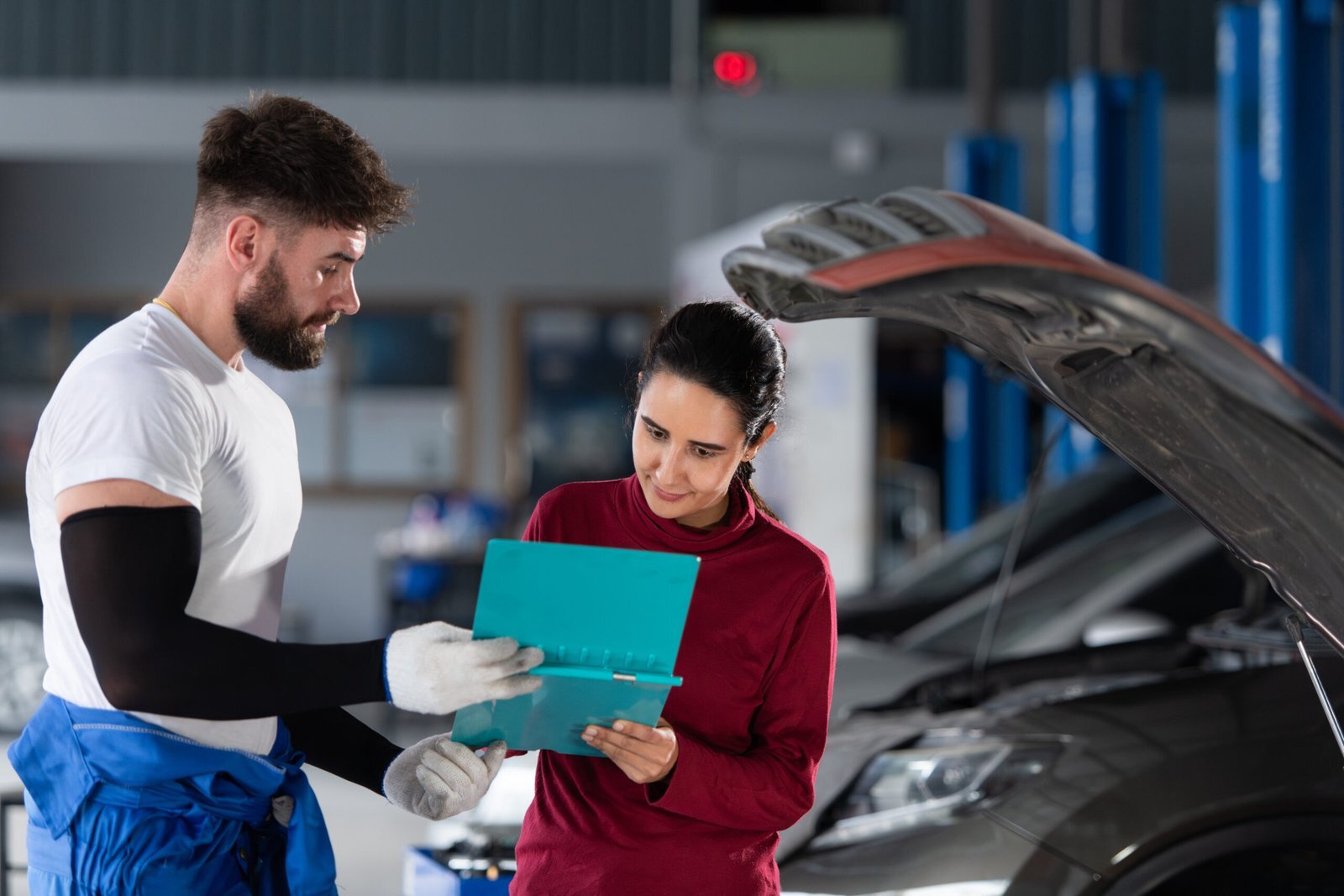 Mécanicien en tenue bleue montrant un document à une cliente dans un garage, capot de voiture ouvert en arrière-plan.