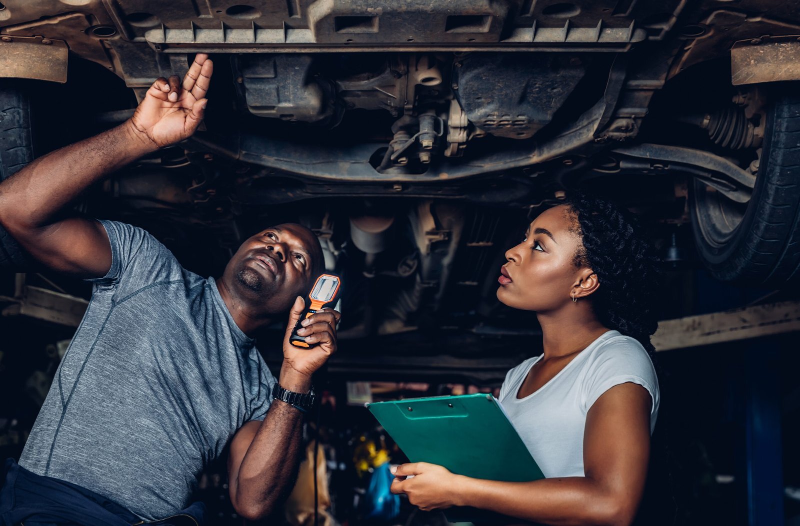 Un expert inspecte minutieusement le dessous d’une voiture pendant qu’une cliente prend des notes, illustrant la transparence et la précision des expertises.